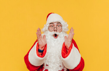 Portrait of shocked Santa Claus on yellow background, looking at camera with open mouth and raised hands in surprise, isolated on yellow background