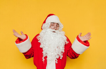Fototapeta na wymiar Puzzled Santa Claus spreads his arms to the side and looks at the camera with a smile on his face, isolated on a yellow background. Christmas and New Year