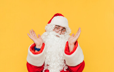 Portrait of a puzzled man in a Santa suit isolated on a yellow background, looking at the camera and confusedly spreads his arms to the side.