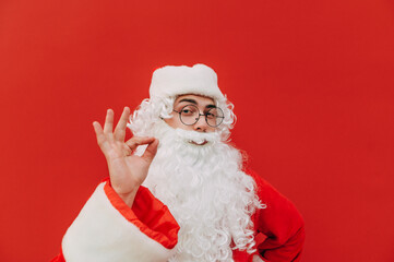 Surprised Santa Claus stands on a background of a red wall and points his finger to the side on the copy space, looking at the camera. Christmas and New Year concept.