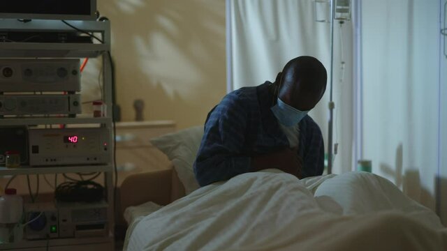 Miserable sick afro-american man suffering chest pain in hospital during night. Lights off. Patient dying from coronavirus infection. Pandemic. No one helps.