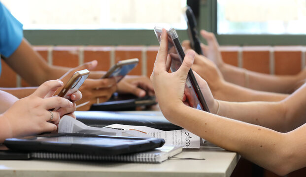 A table of high school students all holding multiple digital devices such as phones and tablets during a class lesson. Contemporary education and connected technology. 