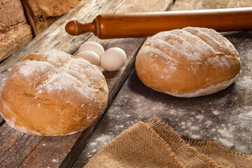 
Homemade round country bread with flour on wooden board with rolling pin, eggs, yeast cloth and rustic cloth with logs in the background