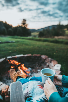 Unrecognizable Woman Enjoying Hot Tea From A Tin Cup In Campsite With Fire Pit. Girl In Folk Blanket By Burning Campfire with mountain landscape with evening sunset sky over the forest and hills.