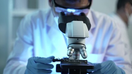 unrecognizable virologist in protective mask, suit and gloves looking through microscope while researching on Covid-19 treatment in laboratory with biohazard sign
