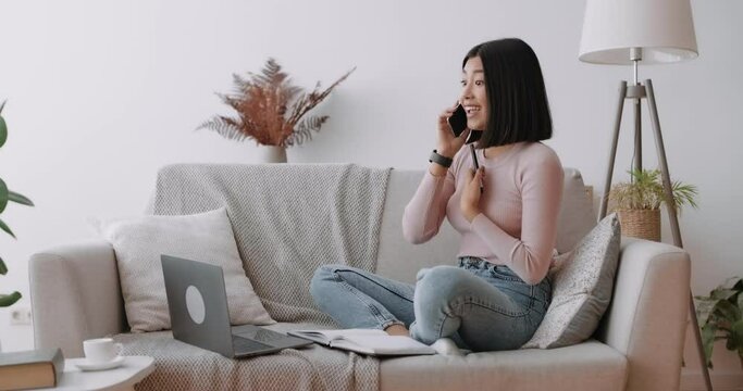 Portrait of young asian woman reacting with surprise during phone call, looking surprised, working with laptop at home