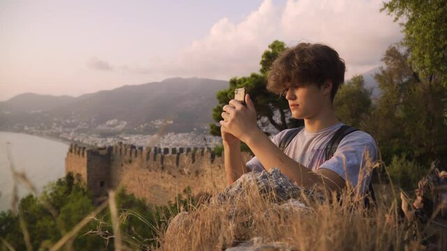 Medium shot of a young man using smartphone at sunset, fortress and mountains in the background, Alanya, Turkey