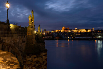 
Charles Bridge on the Vltava river and statues on it and lit street lights and light from lamps at night in the center of Prague in the Czech Republic and there are clouds in the sky