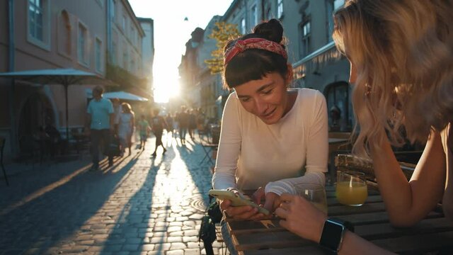 Look at this photo! Two caucasian besties are sitting in the street cafe of an old town and feeling happy communication, laughing and watching something on the phone while enjoying golden sunset