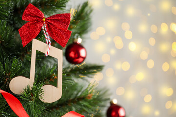 Wooden music note with red bow hanging on Christmas tree against blurred lights. Space for text