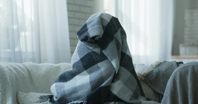 Young depressed woman sitting alone on sofa, hiding from world under plaid at home