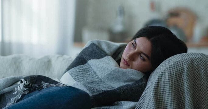 Sad depressed young woman lying alone on sofa wrapped in plaid, feeling tired lonely vulnerable and weak