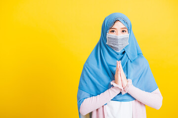 Asian Muslim Arab, Portrait of beautiful young woman Islam religious wear veil hijab and face mask protect quarantines disease coronavirus raising hand praying Eid al Fitr isolated yellow background