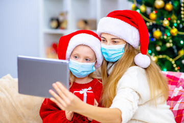 Mom and daughter in medical masks on their face and santa hats on their heads call up with relatives on a tablet computer to wish merry christmas