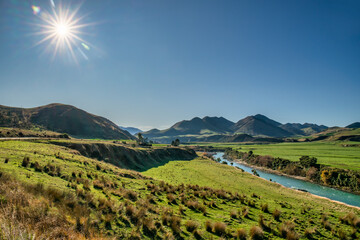 Sun burst over the mountains and the winding Maruia River flowing through sheep grazing on lush farmland pasture in the Lewis Pass