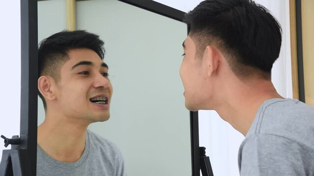 Young Asian man in casual outfit check yourself and smiling with metal braces on teeth in front of mirror for make self confidence