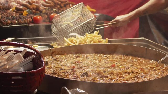 Man throws fries from a metal basket. Cauldron with boiling bigos. Outside. Fast food festival. Lots of fatty junk food.