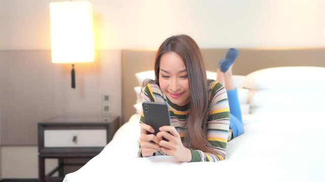 Young Beautiful Asian Woman Laying on Bed, Reading Messages, Articles on Mobile Phone. Social Networking Concept, Full Frame