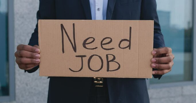 Unemployed black man with poster NEED WORK standing outdoors at office building, close up, tracking shot