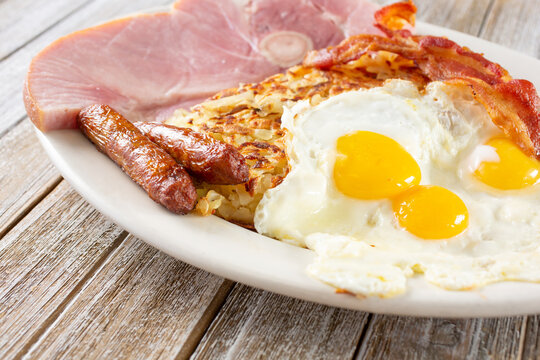 A view of a breakfast plate, featuring sunny side up eggs, sausage links, hash browns, bacon and ham.