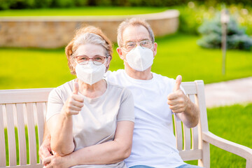 Senior people wearing protective masks sit in a summer park and shows thumbs up gesture. Leisure during the coronavirus epidemic concept