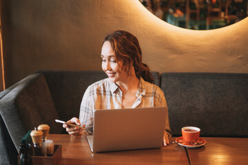 Adult charming brunette woman in plaid shirt working with laptop using mobile phone at the cafe
