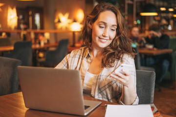 Fototapeta premium Adult charming brunette woman in plaid shirt working with laptop using mobile phone at the cafe