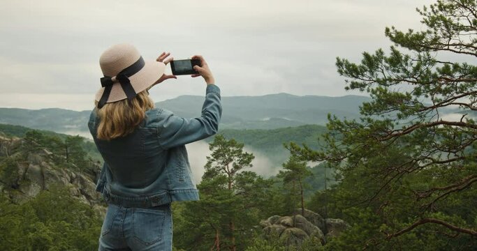 A woman is standing on top of a cliff. She is taking photos of a beautiful mountain landscape. Hiking in the mountains. 4K DCI