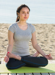 Fototapeta na wymiar Young smiling girl practicing yoga cross-legged in morning by sea