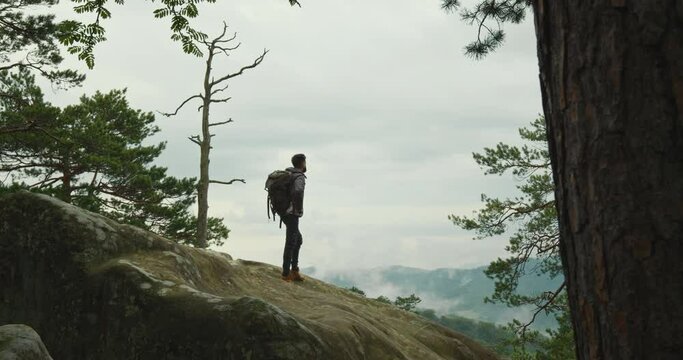 A man is standing on top of a cliff and looking at nature. He is carrying a large backpack. Hiking in the mountains. 4K DCI