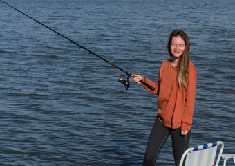 happy girl with closed eyes stands on the shore of the lake with a fishing rod in hand and smiles