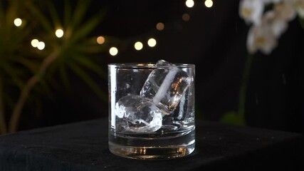 Two ice cubes fly into a fancy glass and a golden drink is poured into it, slow motion - Powered by Adobe