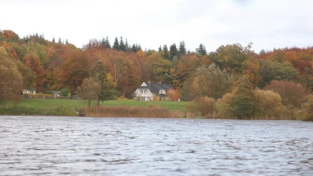 Small waves on a cosy little lake in front of a lone house surrounded by forests painted in autumn colors