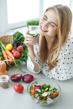 Young woman drinking water near the table with fruits and vegetables in the kitchen. Healthy food, drinks, diet, detox and people concept. High quality photo.