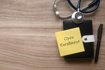 Top view of stethoscope, pen, notebook and yellow paper note written with Open Enrollment on wooden...