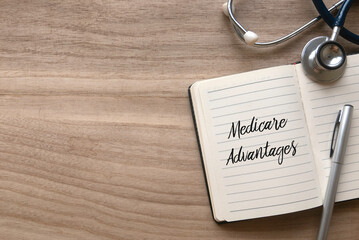 Top view of stethoscope, pen and notebook written with Medicare Advantages on wooden background with copy space.
