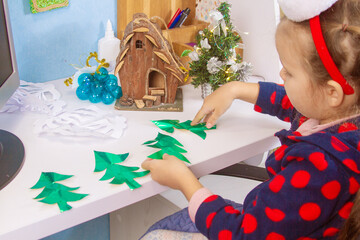 The child prepares for Christmas and New Year, the girl cuts Christmas trees from colored paper,...