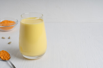 Drinking glass full of golden or turmeric milk which is healthy, healing drink having anti-inflammatory properties served with spoon of curcuma on white wooden background. Image with copy space
