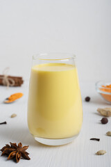 Obraz na płótnie Canvas Drinking glass full of golden or turmeric milk which is healthy, healing drink having anti-inflammatory properties served with anise and cinnamon on white wooden background. Vertical orientation