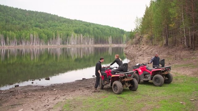 Long shot of men and woman getting on red quad bikes and putting their helmets on after resting by the lake in forest