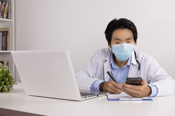 Young Asian Doctor Man in Lab Coat or Gown with Stethoscope Wear Face Mask Writing Report and Using Smartphone and Laptop Computer on Doctor Table in Office
