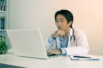 Serious Young Asian Doctor Man in Lab Coat or Gown with Stethoscope Touch Chin and Using Laptop Computer on Doctor Table in Office in Vintage Tone
