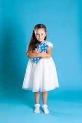 a full-length little Princess in a white dress and snow petal makeup clutches a Christmas gift to her chest, isolated on a blue background.