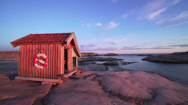 Swedish west coast fishing cabin at sunset. Pink granite rocks at shoreline in purple evening light. Smooth gimbal shot in Sweden Bohuslan ocean coastline. Single red cabin on rocks beautiful nature