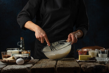 Fototapeta na wymiar The chef in black apron kneads with corolla the dough for cooking waffle on rustic wooden table with ingredients on dark blue background. Frozen motion. Bakery concept.