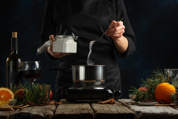 Chef pours sugar into pot with wine fore preparing muled wine on rustic wooden table with festive composition background. Backstage of cooking hot drink with fragrant spices