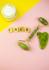 Green Gua Sha Facial Massage Tools. Roller made of green quartz jade on a pink-yellow background. Jar of cream, inscription face in wooden letters.