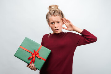 Portrait of puzzled blonde girl 12-15 years old holding gift decorated with ribbon. Studio shot white background