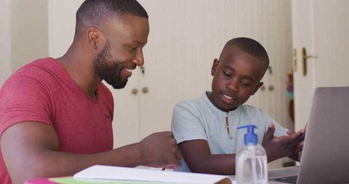 African american father helping son with homework while sitting with laptop and sanitizer bottle on 
