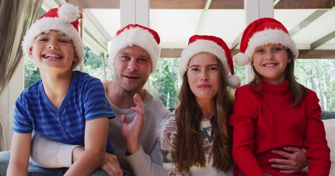 Portrait of caucasian family wearing santa hats smiling and waving looking at the camera while sitti